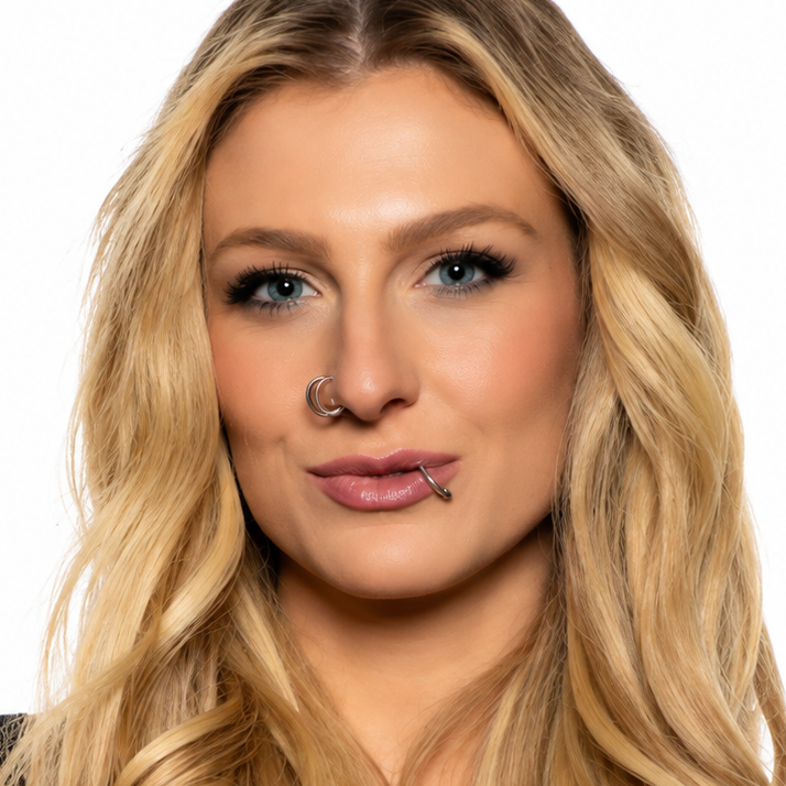 A woman with long, wavy blonde hair and nose jewelry, wearing a black top against a plain white background.