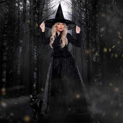 A woman wearing a black witch costume with a tall pointed hat stands in a dark misty forest, holding the brim of the hat with both hands. A small black cat is at her side.