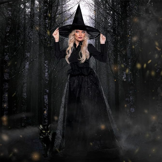 A woman wearing a black witch costume with a tall pointed hat stands in a dark misty forest, holding the brim of the hat with both hands. A small black cat is at her side.