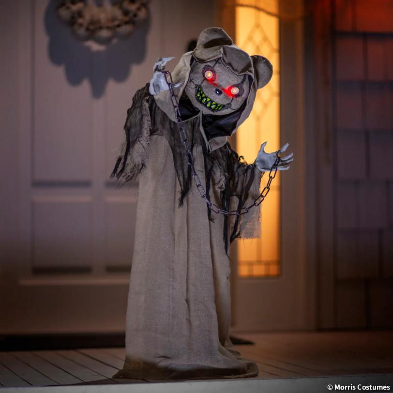 A spooky Halloween decoration shaped like a ragged ghost bear in a tattered robe, with glowing red eyes, green teeth, and a chain around its neck, posed in front of a house door and window.