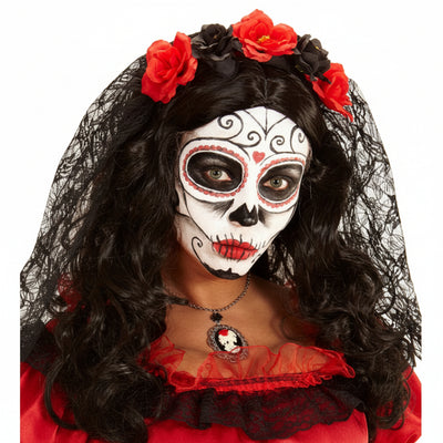 A person wearing Day of the Dead style face makeup with white, black, and red skull design, long black wavy hair, red flowers in the hair, and a black lace veil. The image is tightly cropped from the chest up against a white background.