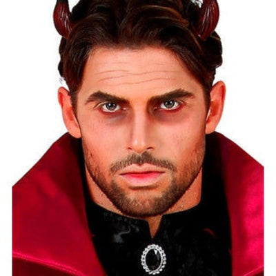 A close-up of a man wearing a vampire costume accessory set, featuring dark brown horns on his head, dramatic red eye makeup, a dark beard, a black collar with a jeweled silver emblem, and a red satin cape against a white background.