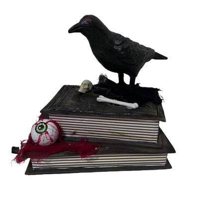 21cm animated crow Halloween decoration with black raven perched on stacked dark spell books with miniature skull, bone and decorative eyeball prop on tattered red fabric