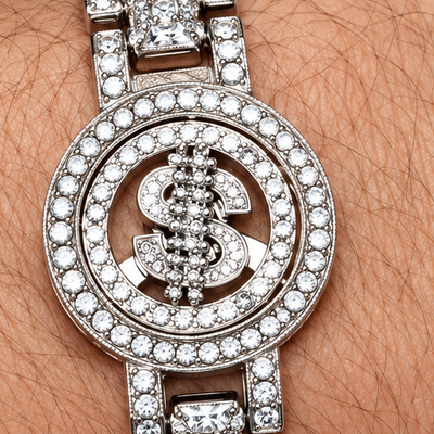 A close-up of a wrist wearing a silver-toned watch heavily embellished with clear crystals, featuring a circular crystal-encrusted face with a dollar-sign motif in the center.